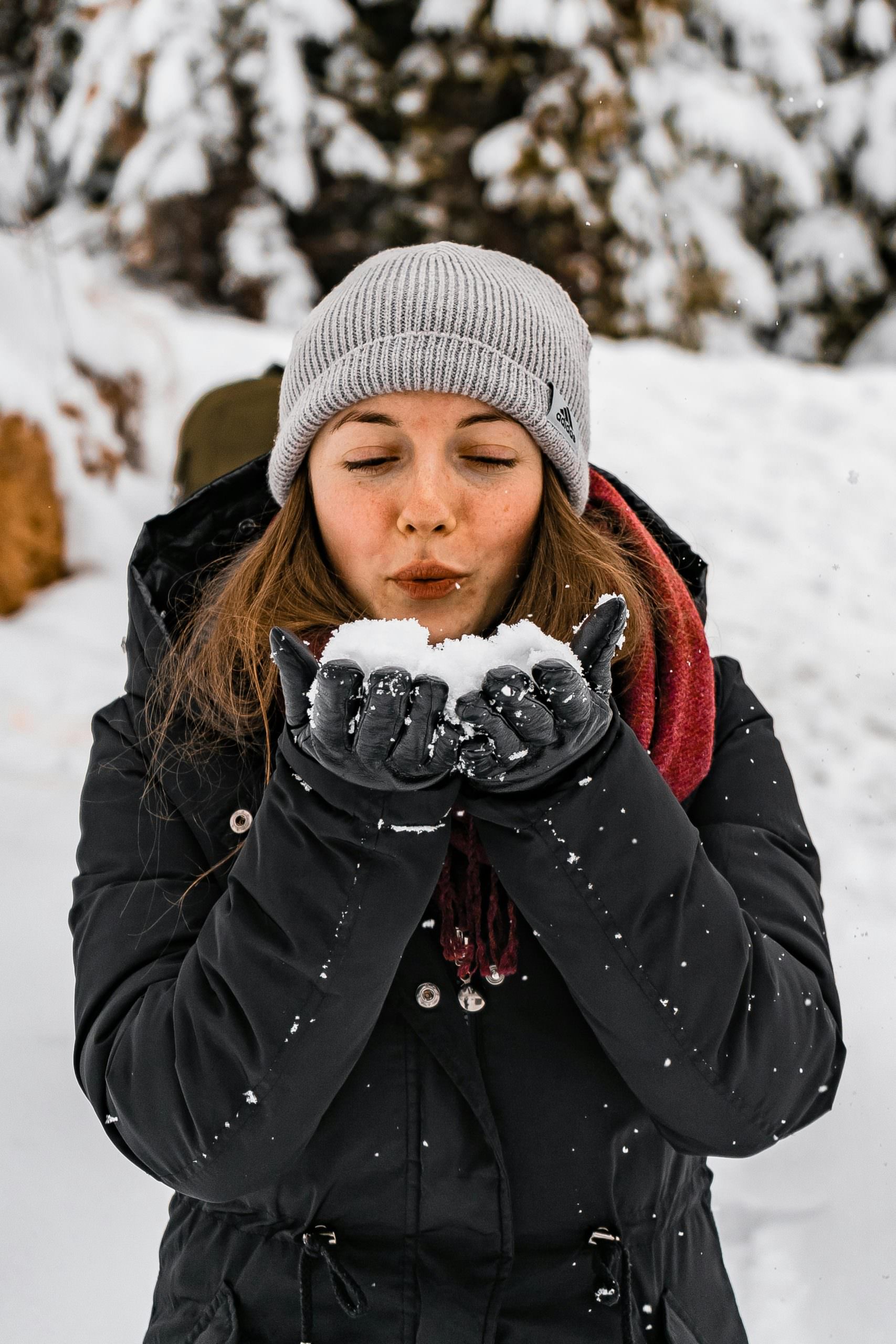 Donna vestita per il freddo con giacca invernale, guanti, sciarpa e berretto di lana, mentre soffia sulla neve tra le mani in un paesaggio innevato. Outfit perfetto per affrontare meno 10 gradi con calore e comfort.