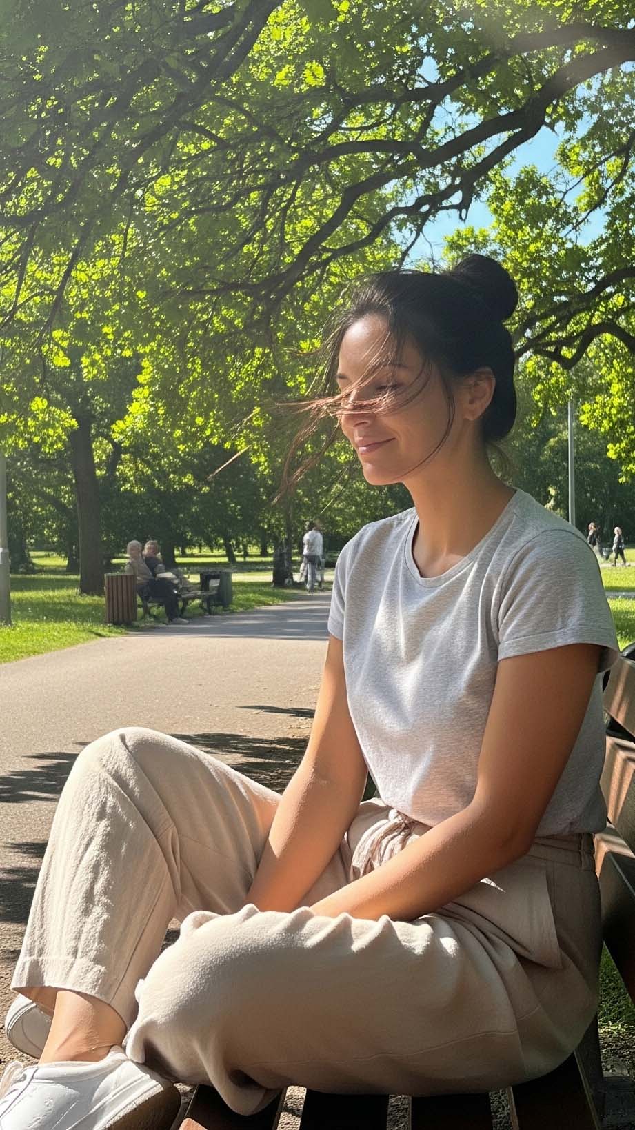 Una donna rilassata seduta su una panchina al parco in una giornata di sole, indossa un outfit ideale per 25 gradi: pantaloni chiari in lino e una t-shirt grigia in cotone