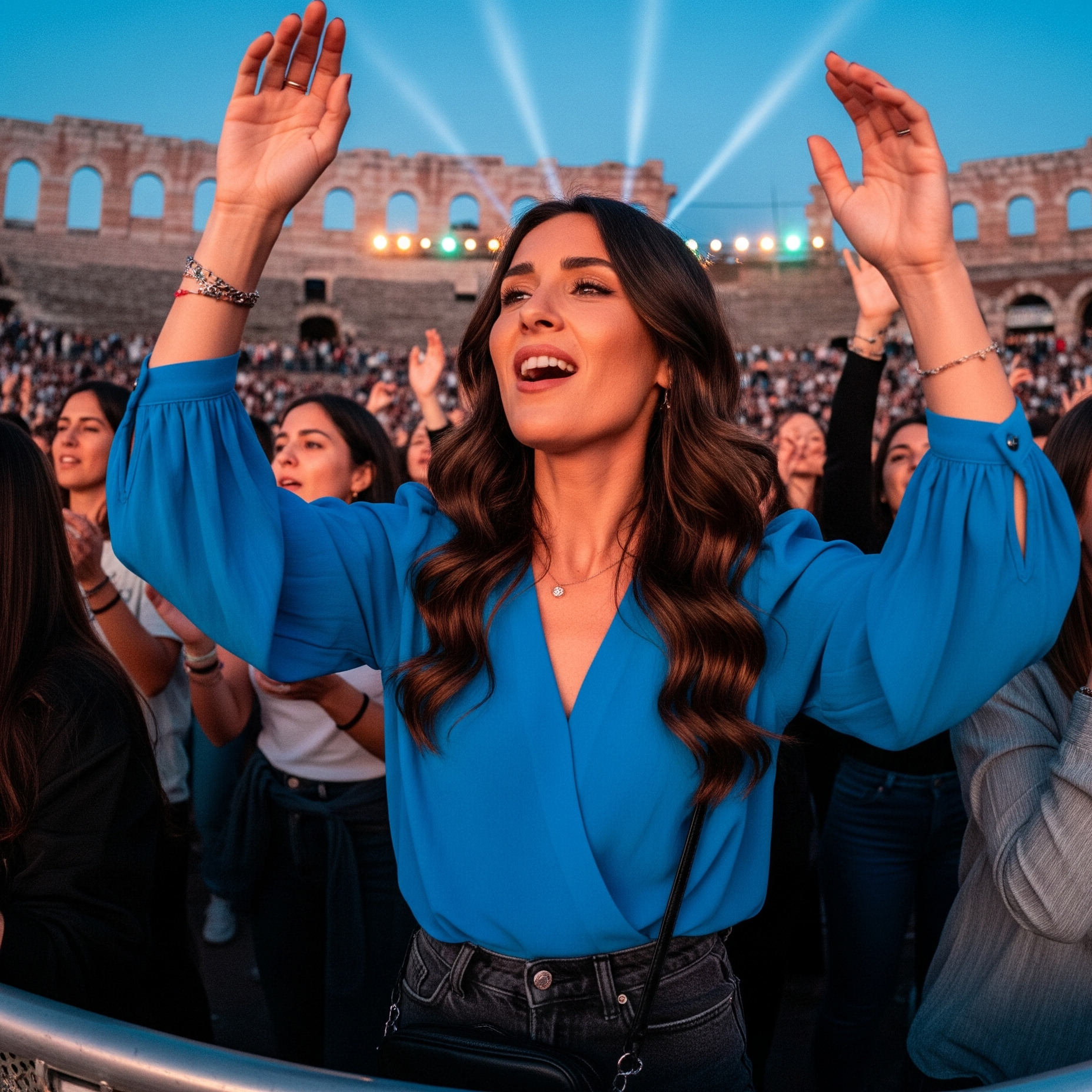 Una ragazza entusiasta con le mani al cielo, che indossa una blusa blu con maniche a sbuffo e jeans, durante un concerto all'Arena di Verona.


