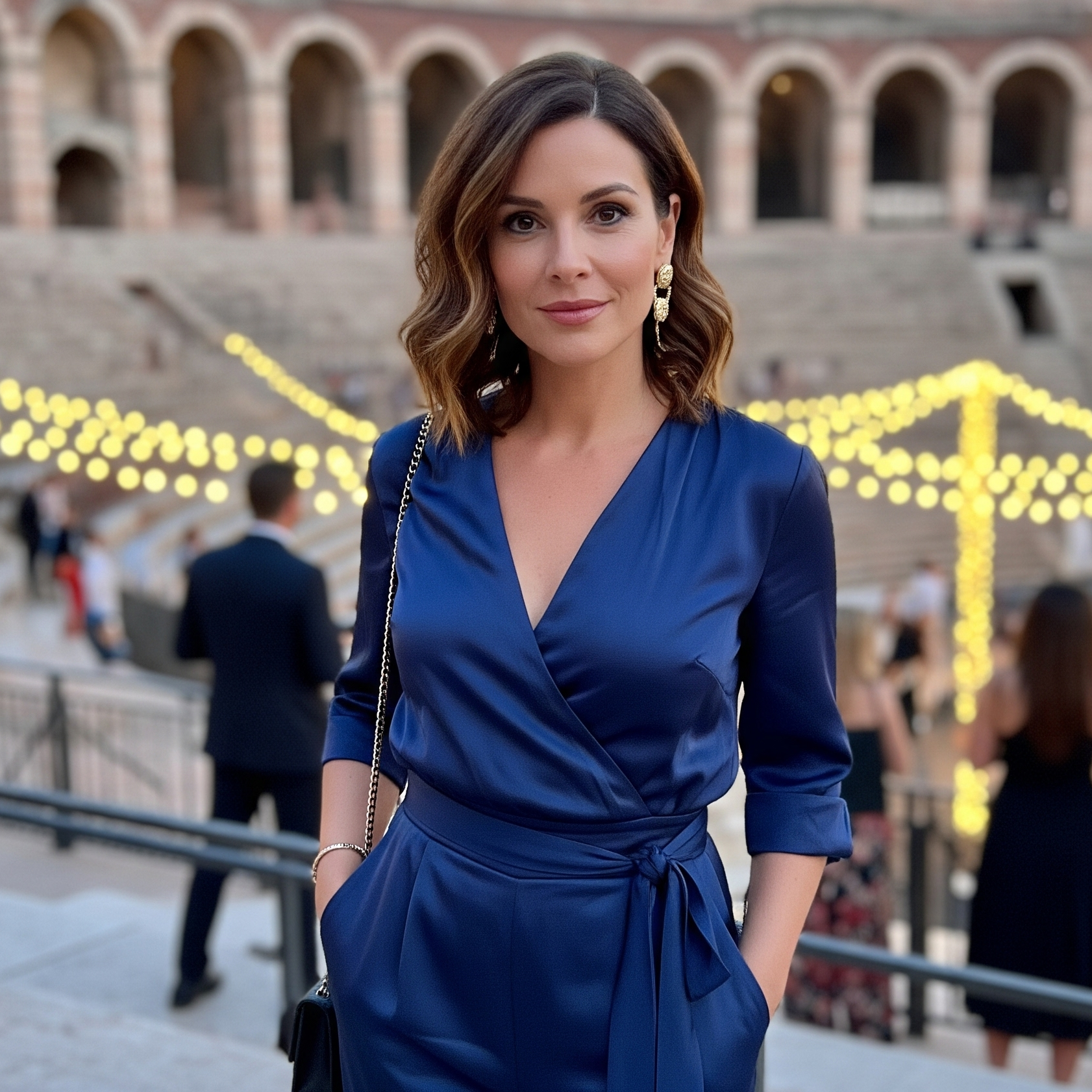 Donna con i capelli castani indossa una jumpsuit elegante blu scuro mentre si trova sulle gradinate dell'Arena di Verona durante un evento serale.

