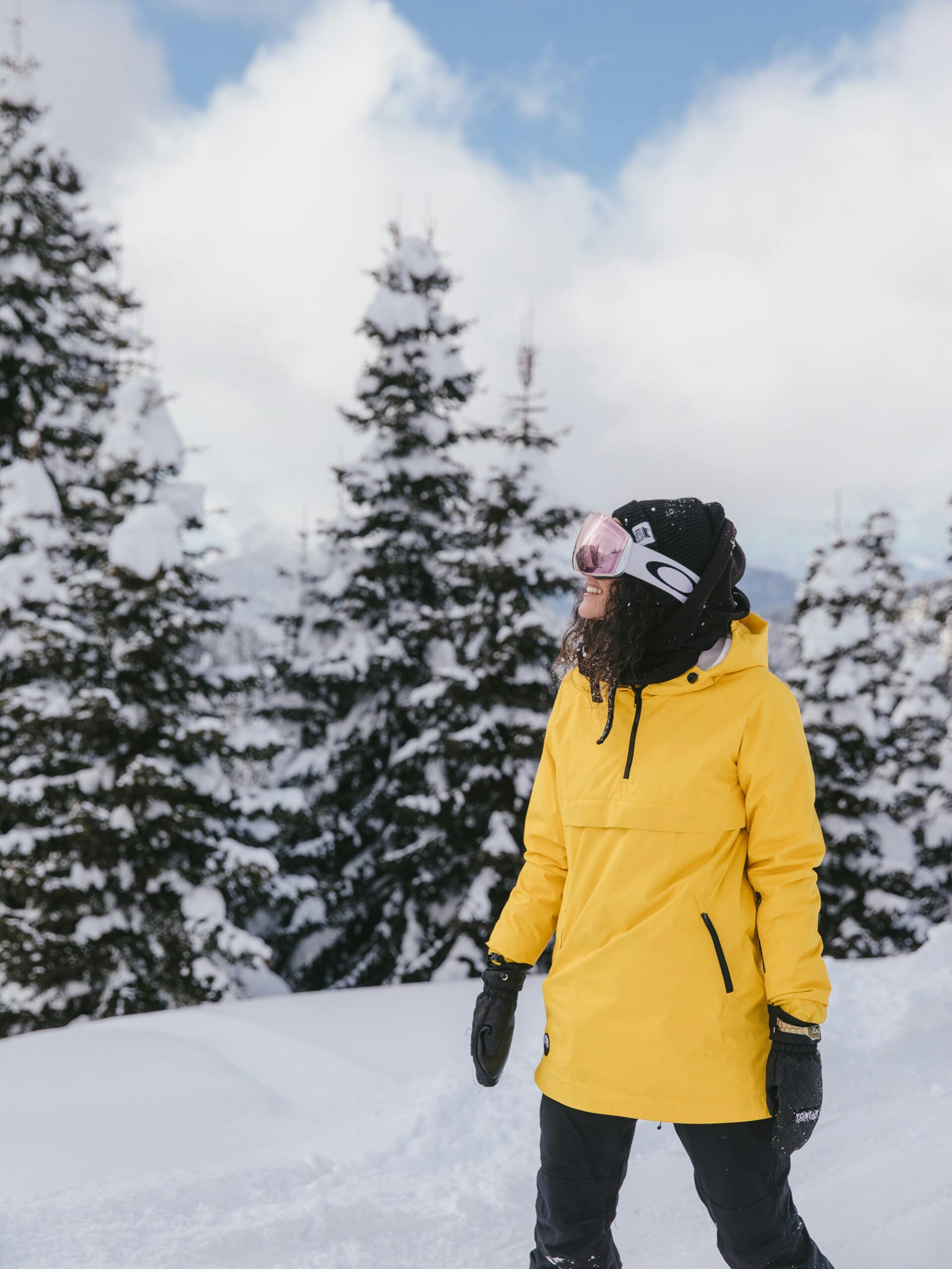 Donna con giacca tecnica da sci gialla, pantaloni neri e maschera da neve in un paesaggio montano innevato