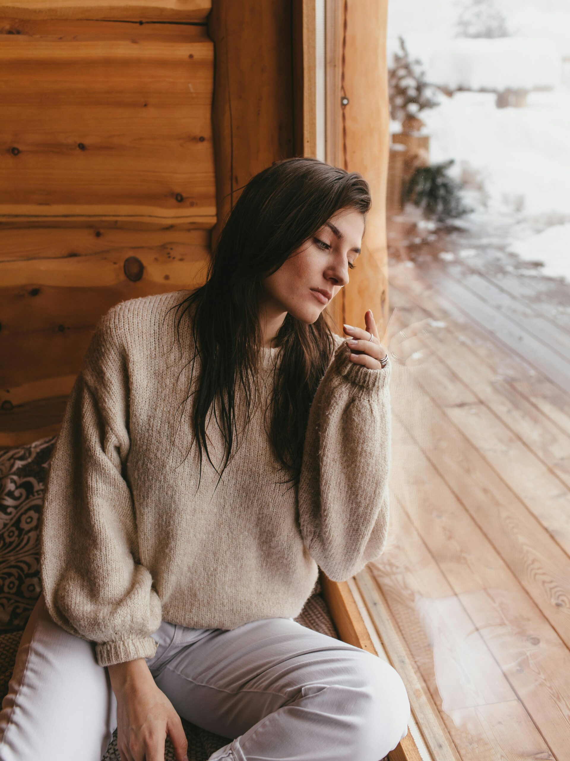 Come vestirsi con temperature sotto lo zero? 3 Donna seduta vicino a una finestra in una cabina di legno, indossa un maglione in lana beige e pantaloni chiari, outfit caldo e accogliente per l’inverno con neve all’esterno.