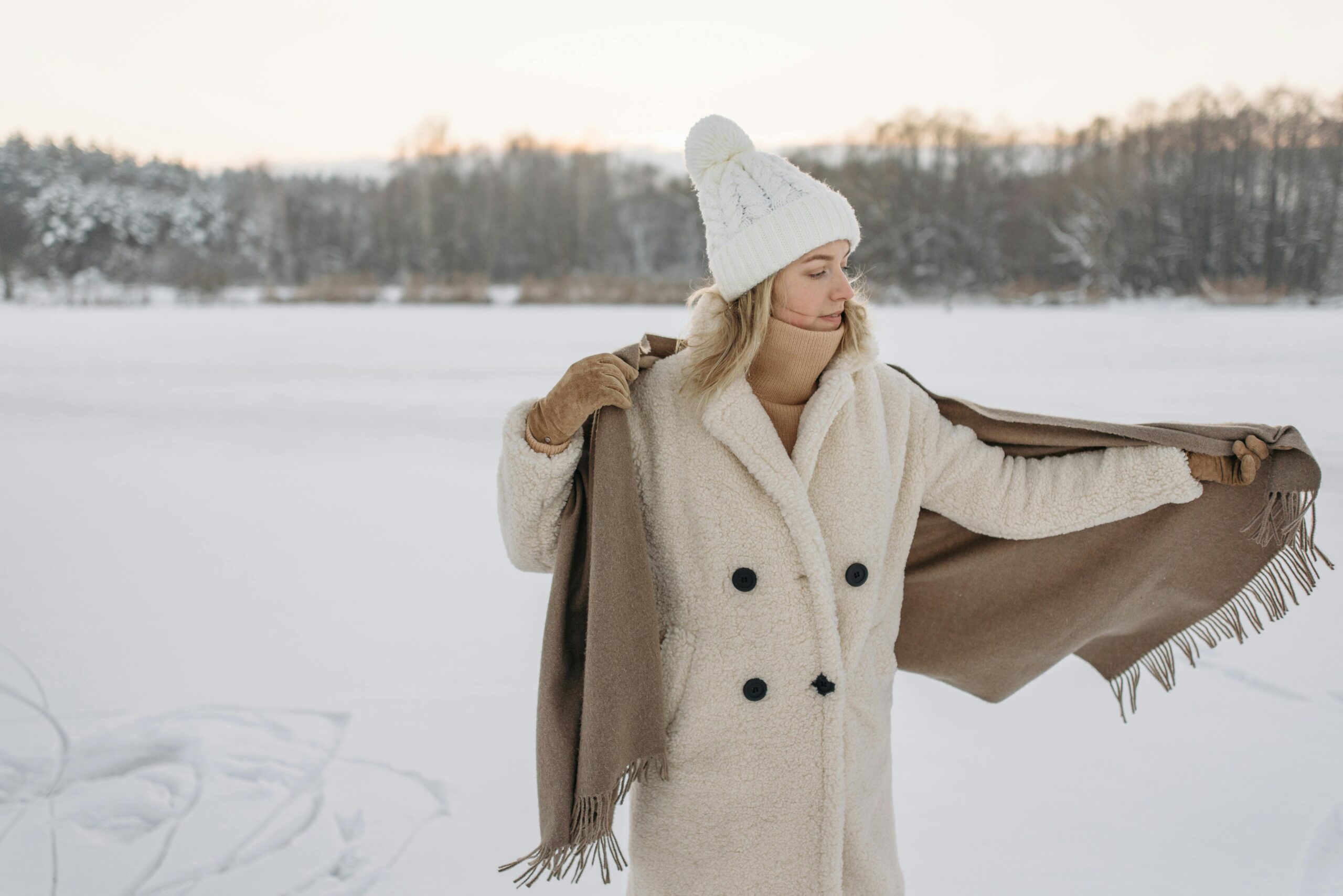 Come vestirsi con temperature sotto lo zero? 6 Donna all’aperto sulla neve che indossa un cappotto in pelliccia sintetica color crema, berretto bianco e guanti, mentre sistema una sciarpa calda; outfit elegante e termico per temperature sotto lo zero.