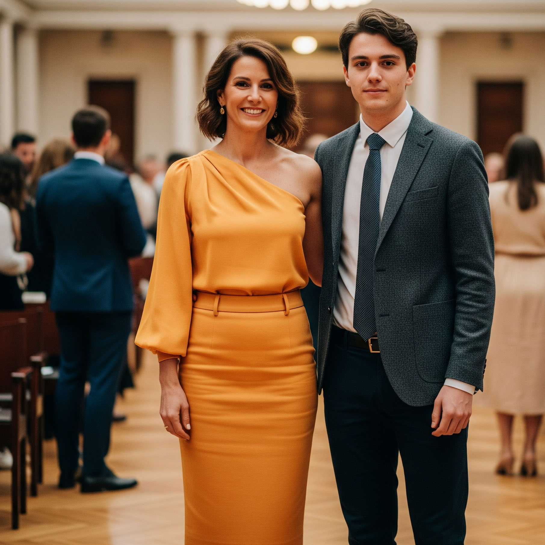 Outfit laurea per la mamma: donna elegante con completo moderno composto da gonna midi e top monospalla giallo ocra, posa con il figlio laureato.

