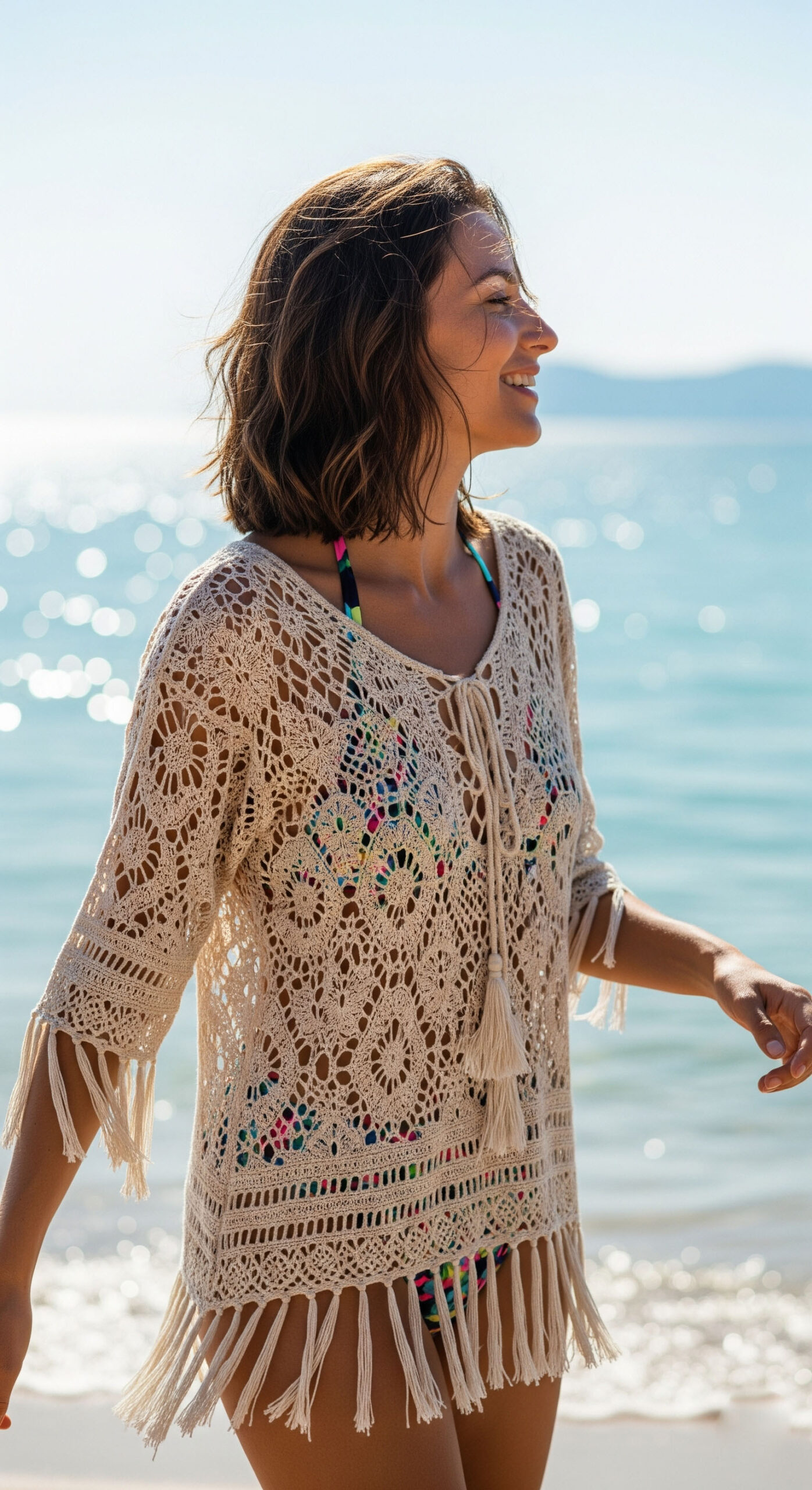 Donna sorridente in spiaggia con un look da mare per agosto, indossa un copricostume crochet color crema con frange sopra il bikini.

