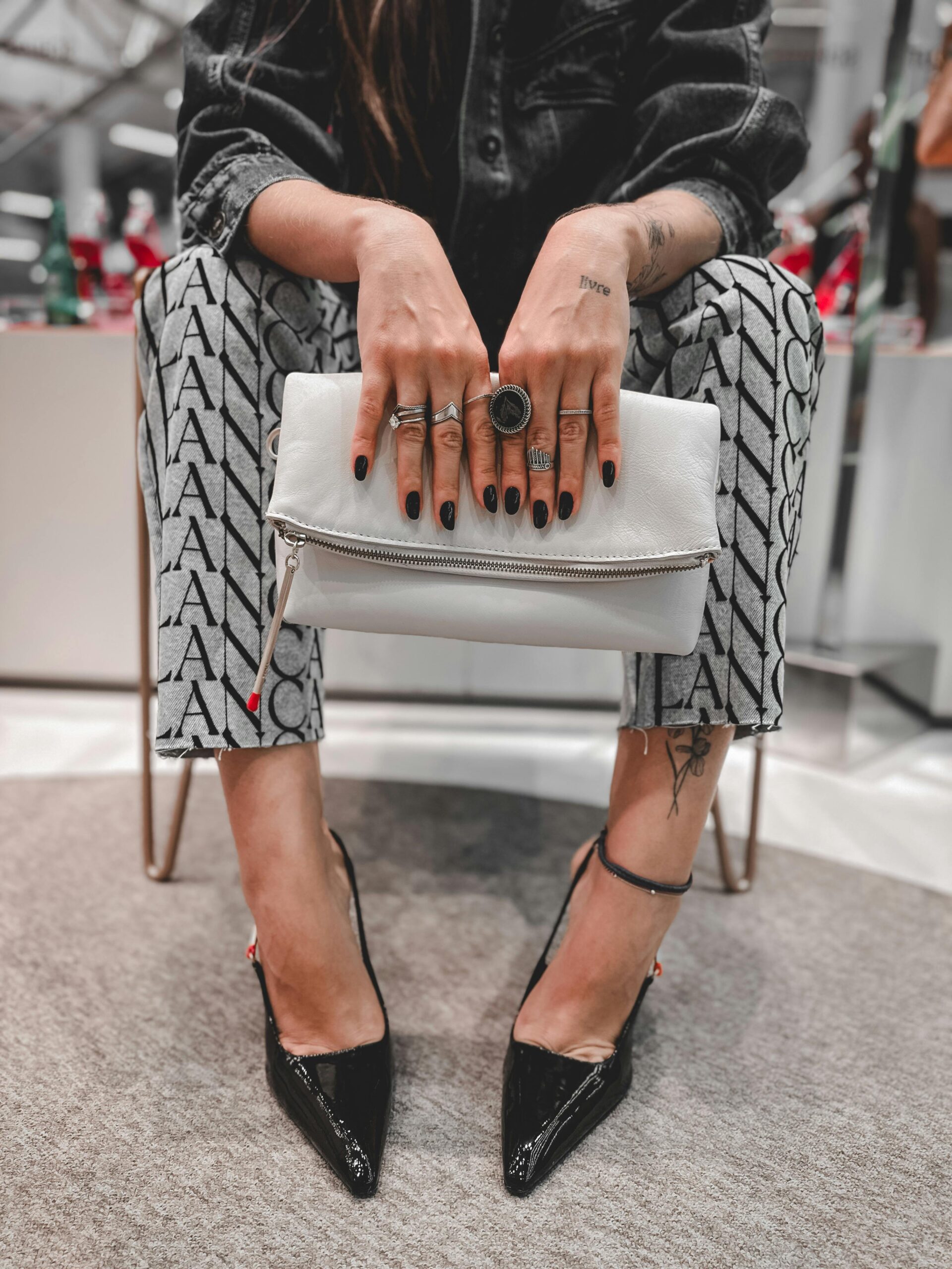 7 colori di smalto che stanno bene su tutto da Provare Ora 6 Donna seduta con manicure nero lucido, anelli silver, borsa bianca e tacchi neri a punta; outfit streetwear elegante con pantaloni stampati.