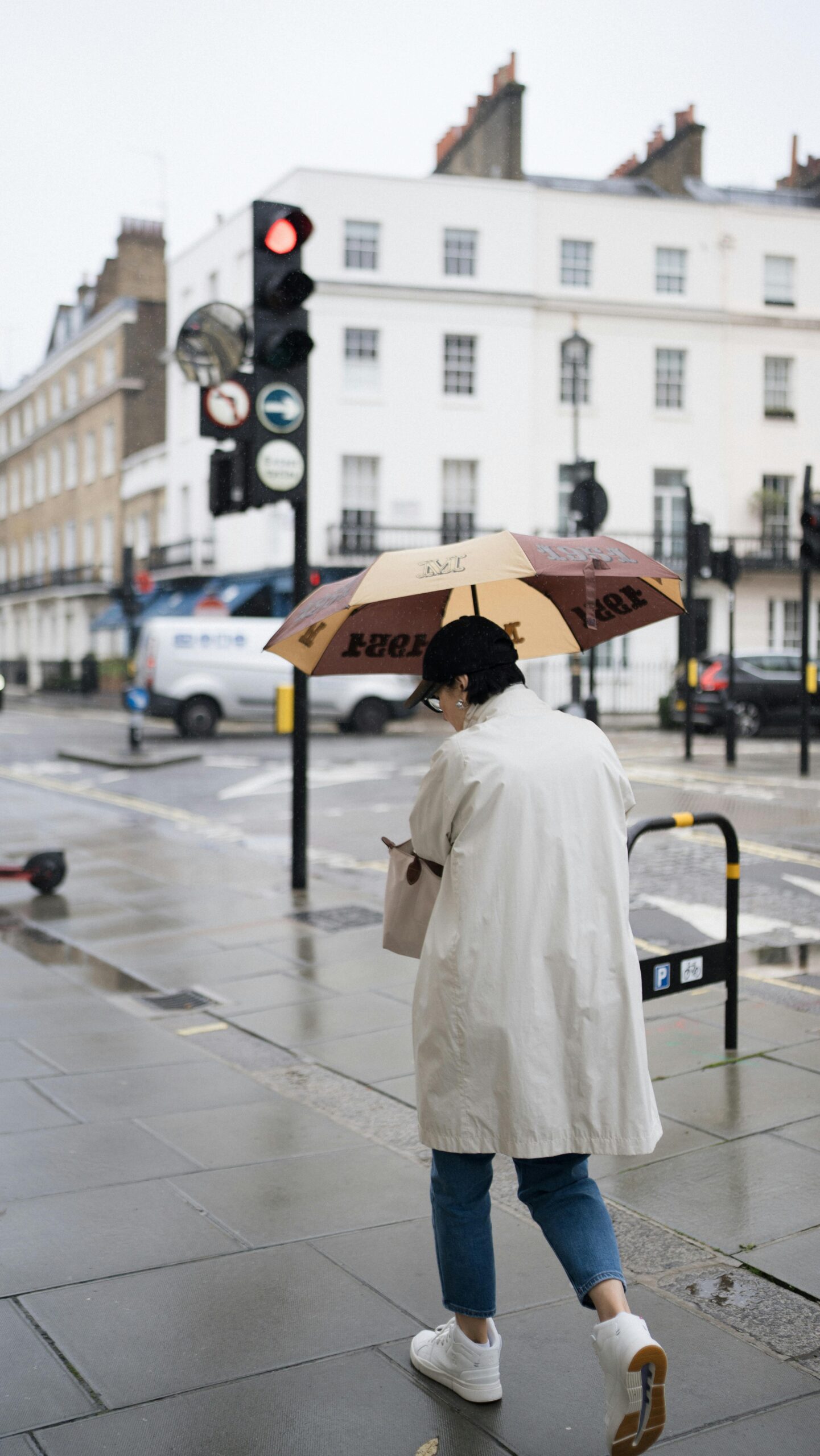 Donna a Londra in un giorno di pioggia con trench chiaro, jeans e sneakers bianche sotto l’ombrello, esempio di outfit casual per come vestirsi a Londra quando piove