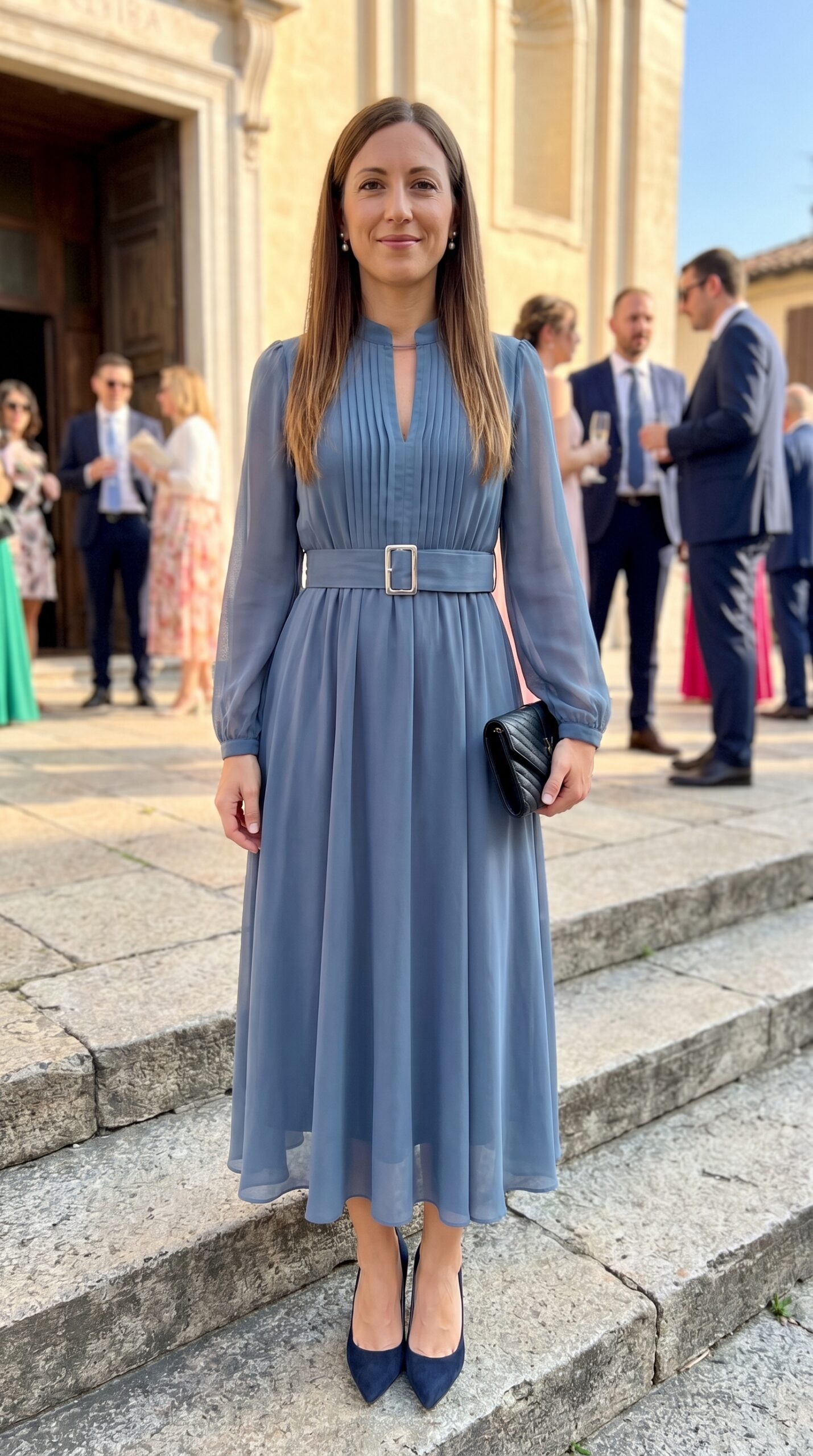 Invitata elegante indossa un abito midi plissettato color azzurro carta da zucchero con maniche velate e accessori scuri.