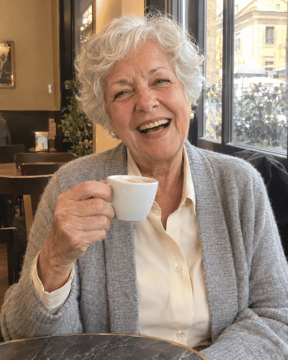 Come Vestirsi a 70 Anni: Guida Completa e Consigli Pratici 8 Donna di 70 anni con cardigan grigio perla e camicia crema al bar con un caffè