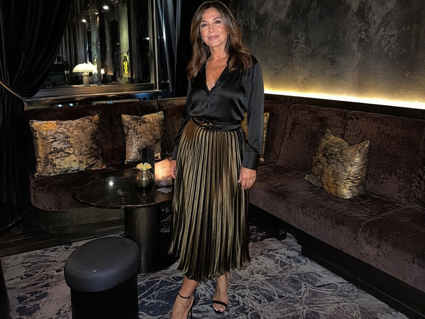 Donna di 60 anni giovanile con gonna midi plissettata color bronzo e camicetta in seta nera in un lounge elegante di sera