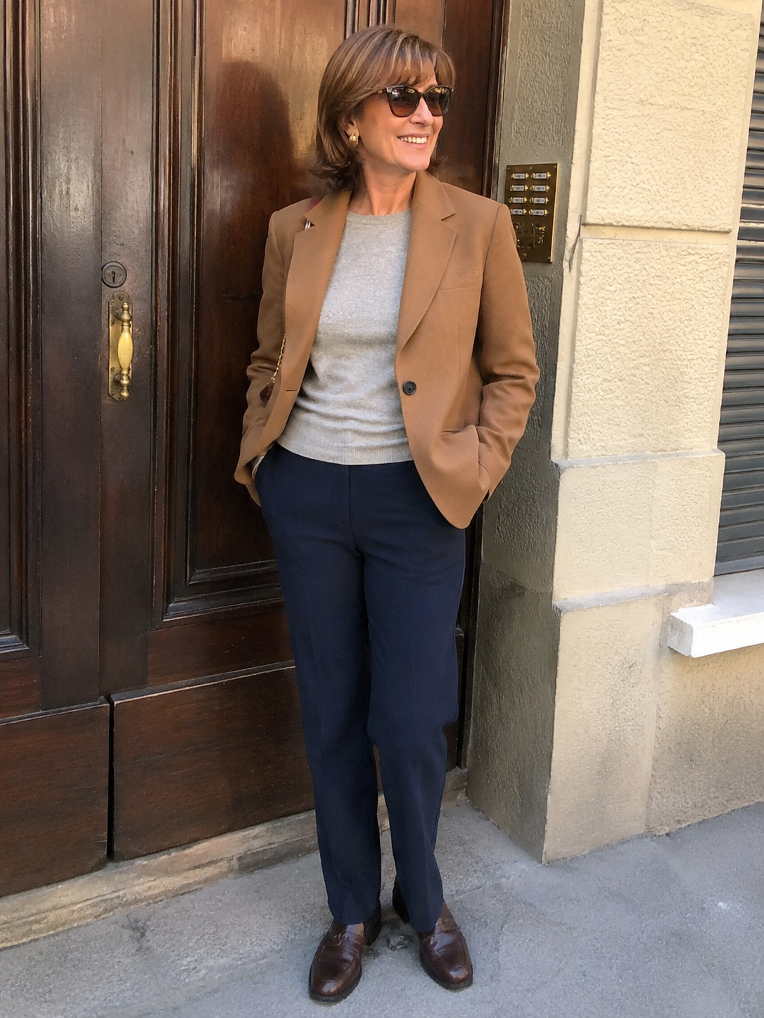 Donna di circa 60 anni con pantalone dritto blu, maglia fine grigia, blazer cammello e mocassini, outfit elegante da giorno