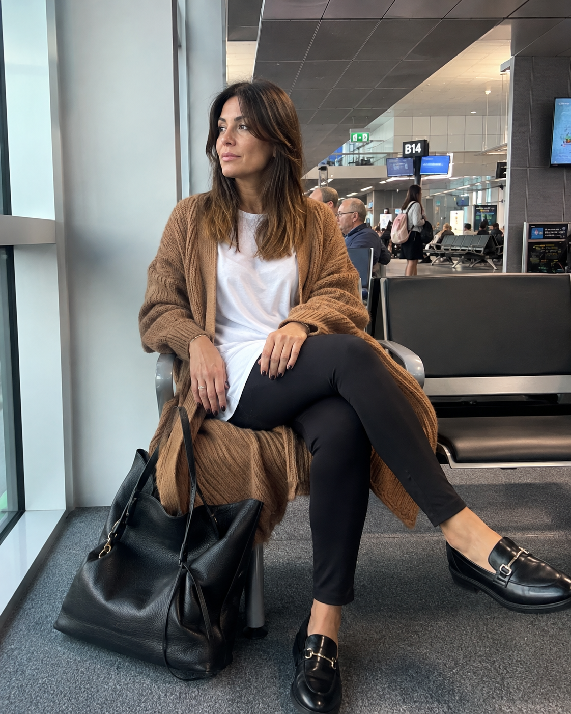 Donna di 40 anni seduta nella sala d'attesa di un aeroporto con leggings neri, t-shirt bianca lunga, cardigan cammello, mocassini e borsa shopper