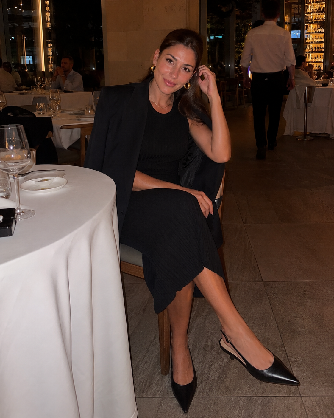 Donna di 40 anni con vestito nero midi in maglia a costine, blazer, slingback basse e orecchini dorati durante una cena al ristorante