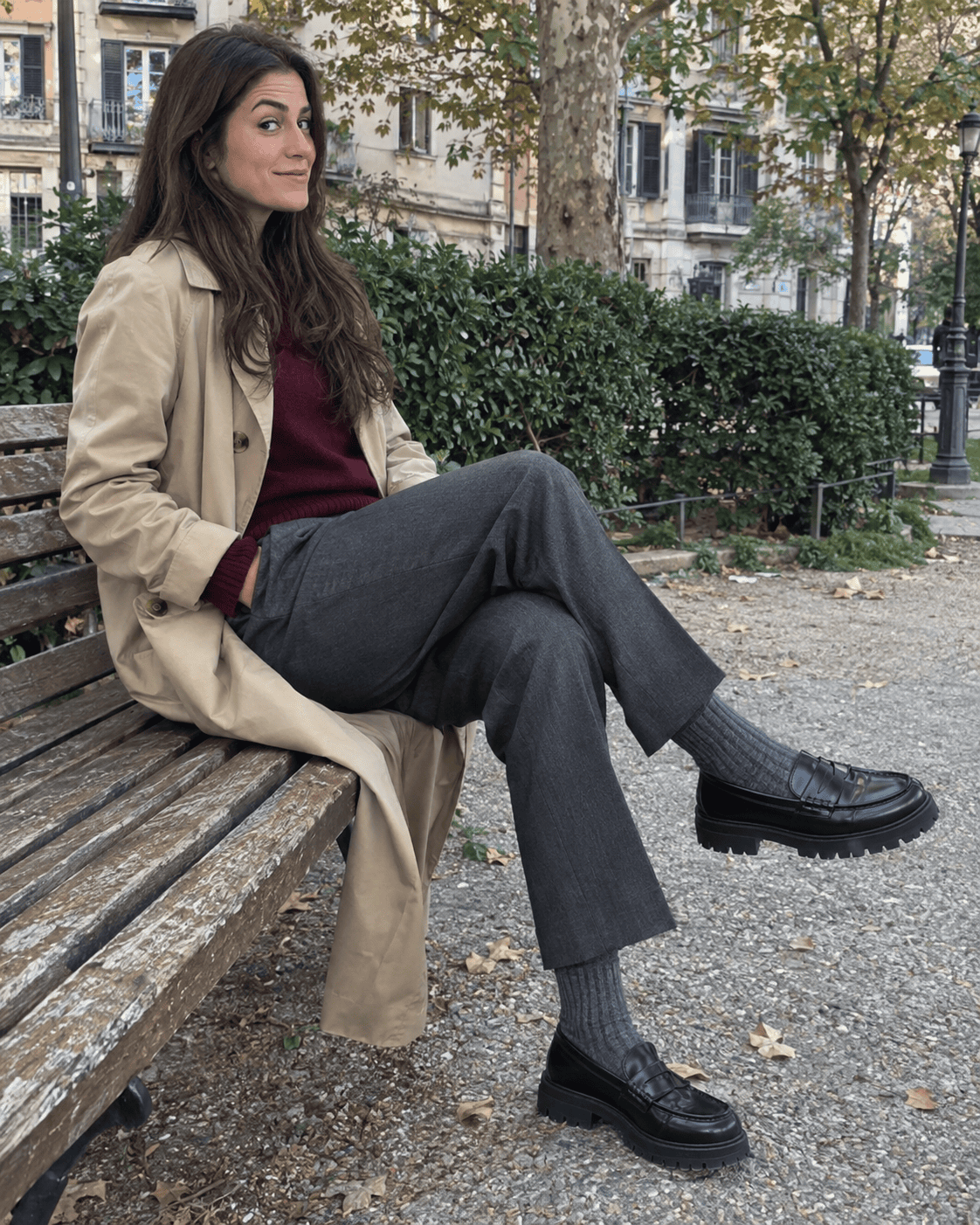 Mocassini neri con calzino grigio a costine, pantaloni grigi, maglione bordeaux e trench beige
