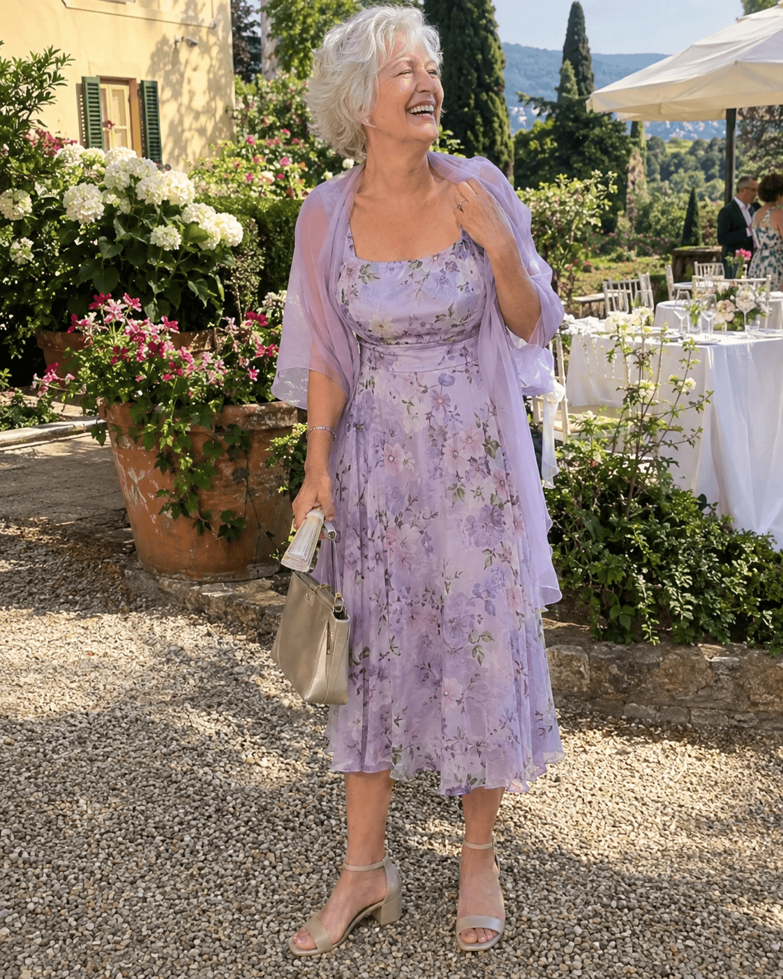 Nonna romantica al battesimo con abito midi in chiffon lilla a fiori, coprispalle in tulle, sandali con tacco quadrato e ventaglio nel giardino di una villa