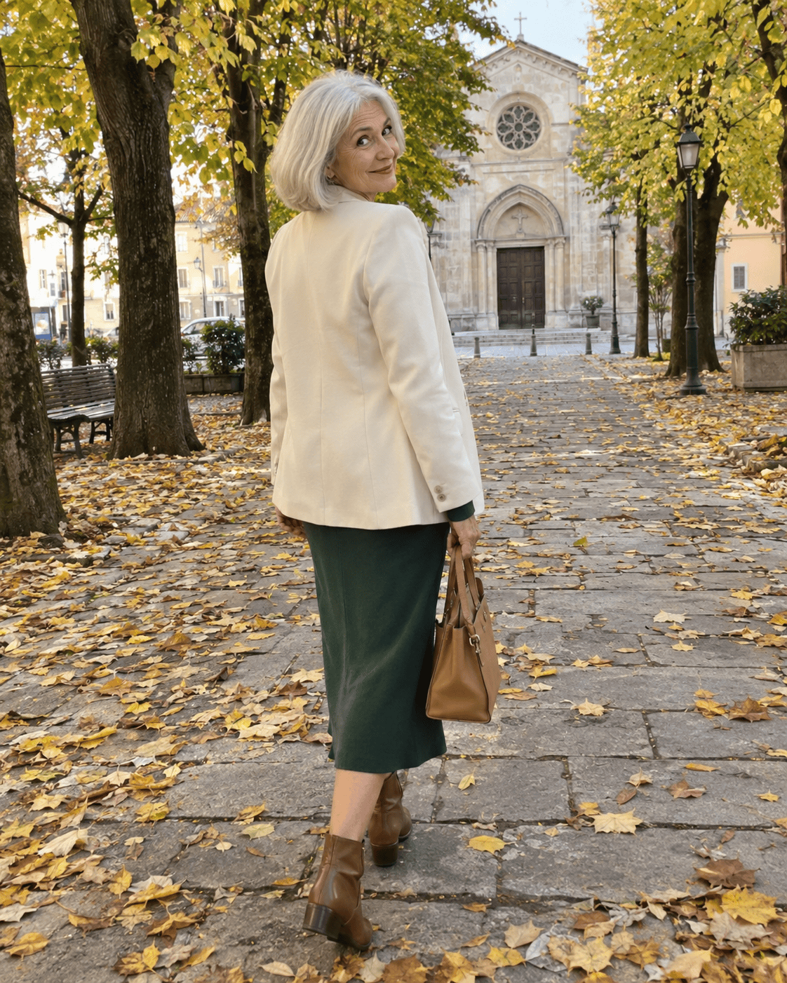 Nonna al battesimo in autunno con abito verde bosco, blazer crema, stivaletti in pelle e borsa cammello sul viale davanti alla chiesa