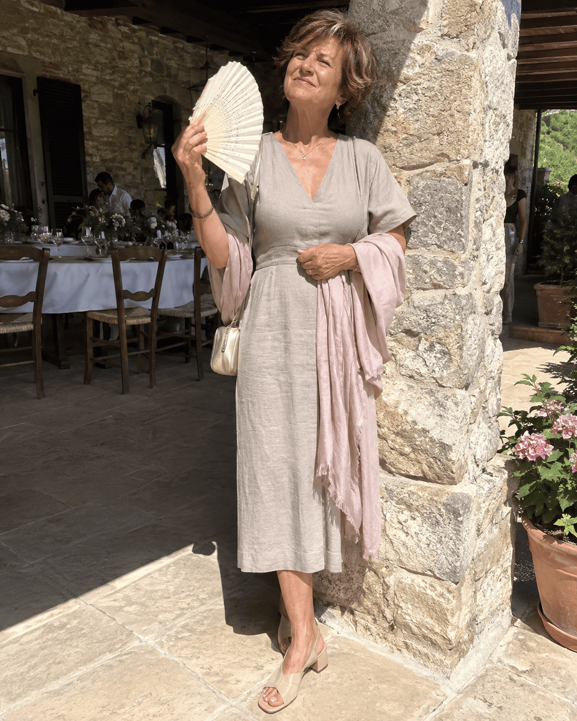 Nonna al battesimo in estate con abito midi in lino color tortora, scialle rosa cipria, sandali nude con tacco basso e ventaglio avorio sotto il portico di una casa di campagna