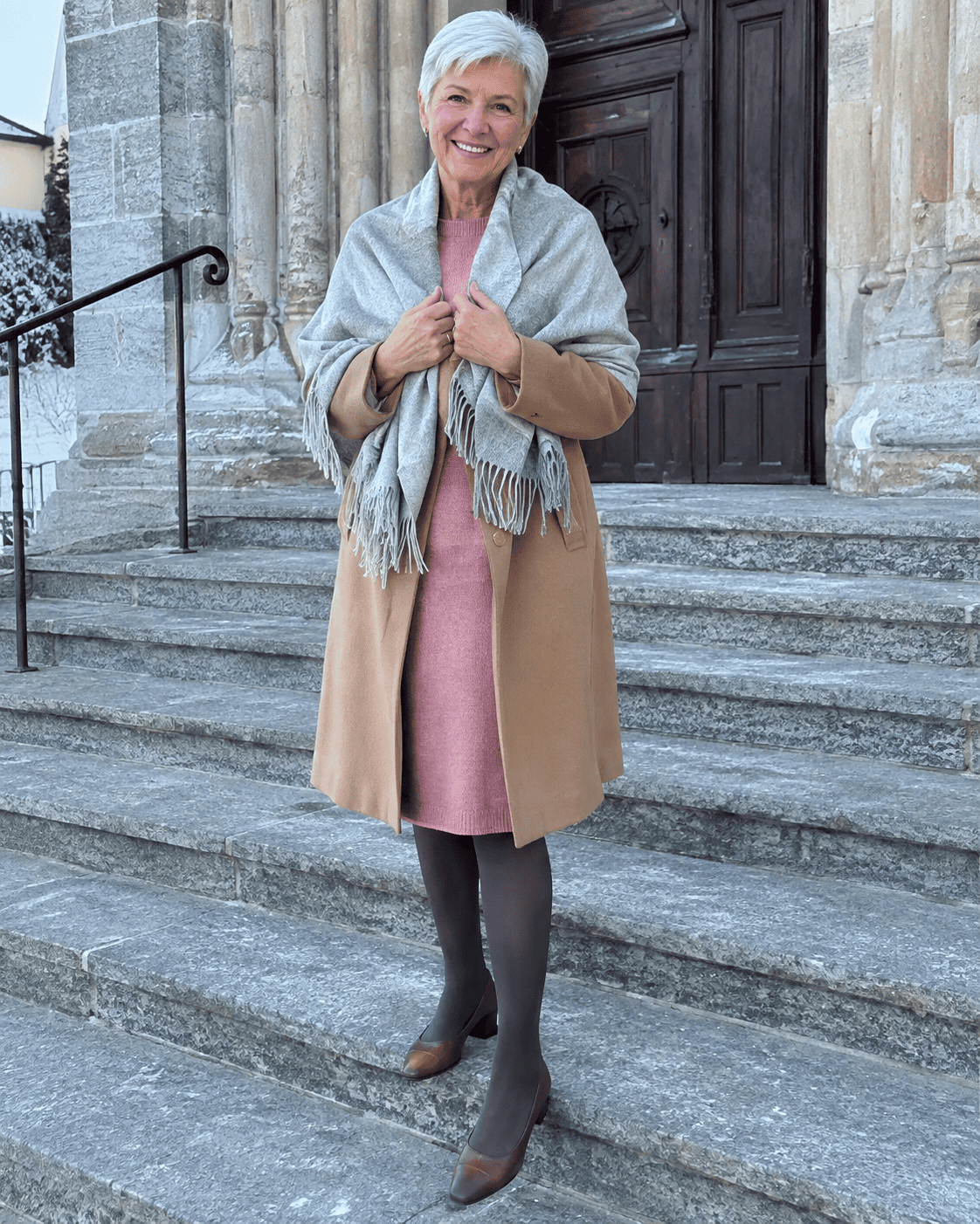 Nonna al battesimo in inverno con cappotto cammello, abito in lana rosa antico, stola in cashmere grigio perla, collant coprenti e décolleté marroni sulla scalinata della chiesa