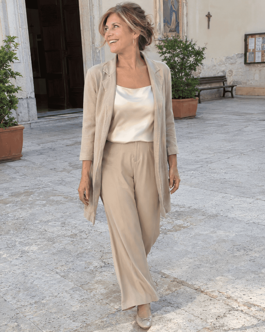 Nonna moderna al battesimo con pantaloni palazzo color sabbia, top in seta avorio, giacca lunga leggera e ballerine beige nel cortile della chiesa