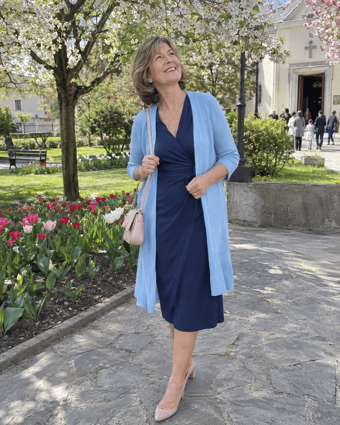 Nonna al battesimo in primavera con abito a portafoglio blu navy, cardigan lungo azzurro polvere, décolleté nude e borsa a tracolla cipria nel parco fiorito davanti alla chiesa