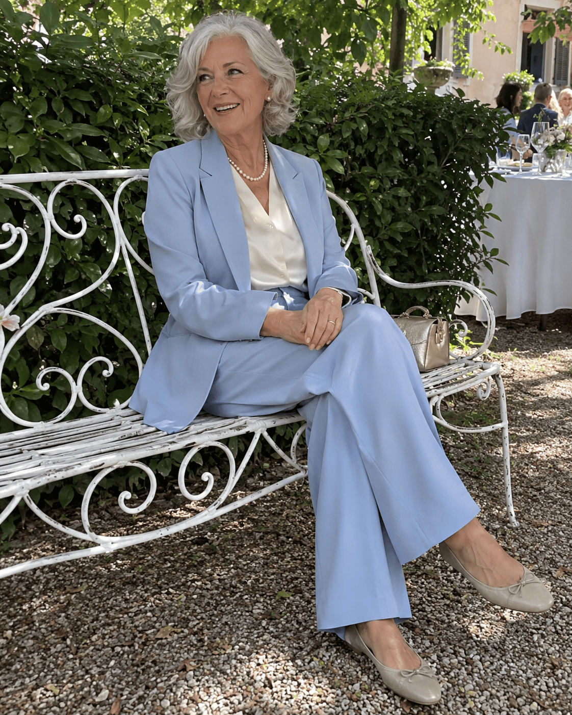 Nonna al battesimo con tailleur pantalone azzurro polvere, camicetta di seta avorio, collana di perle e ballerine beige, seduta su una panca nel giardino della villa