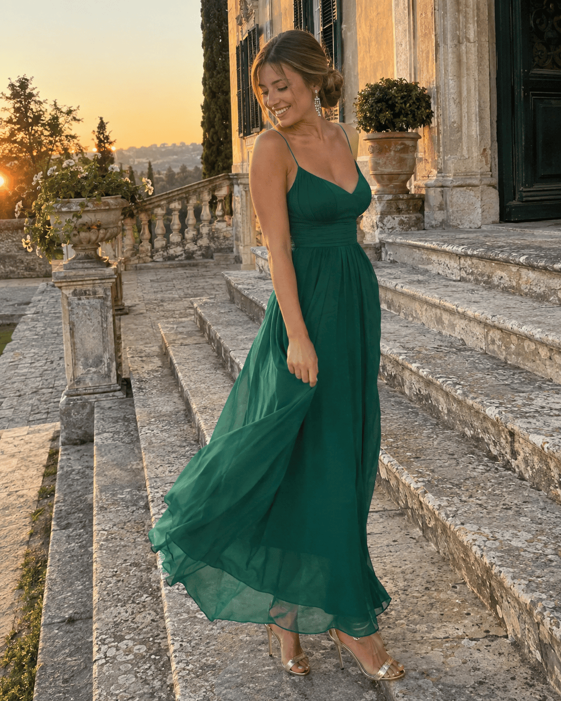 Donna in abito lungo a trapezio in chiffon verde smeraldo con spalline sottili, sandali dorati e orecchini pendenti sulla scalinata di una villa storica al tramonto