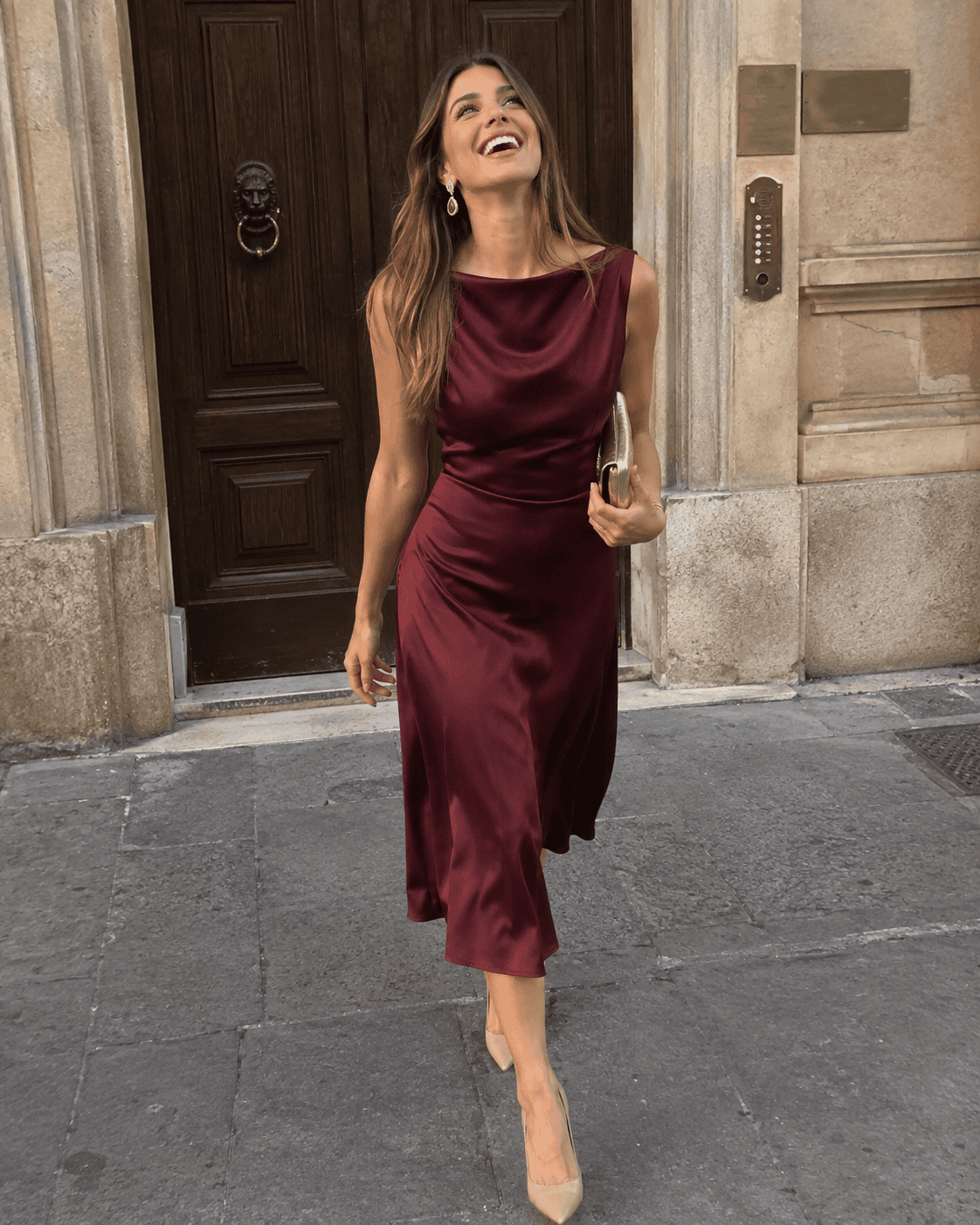 Donna sorridente in abito midi in seta bordeaux con scollo a barchetta, décolleté nude con tacco alto e clutch dorata davanti al portone di un palazzo storico