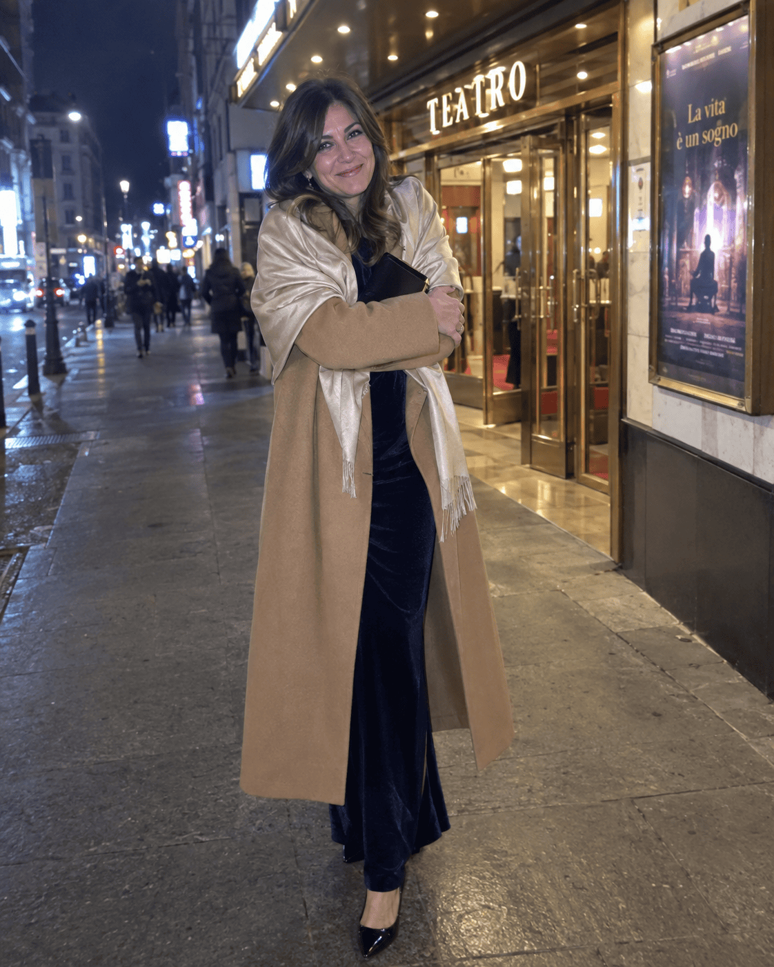 Donna con cappotto lungo color cammello e stola in seta champagne sopra un abito lungo in velluto blu notte e décolleté nere davanti all'ingresso di un teatro di sera