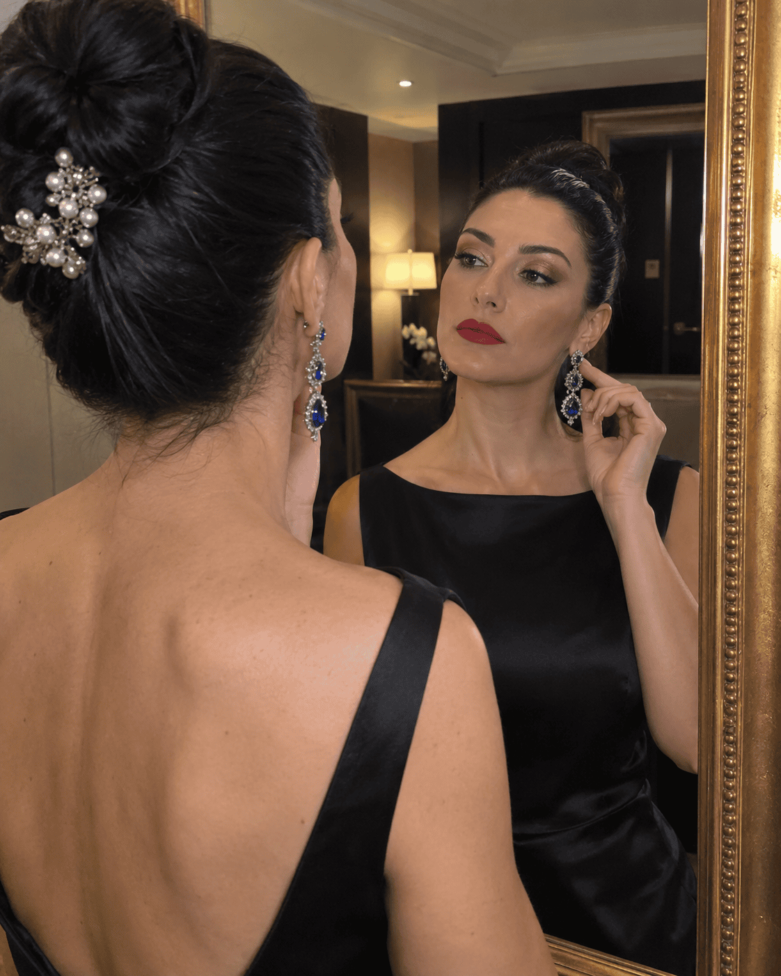 Donna allo specchio con chignon alto, fermaglio gioiello con perle, orecchini chandelier con pietre blu, rossetto rosso e trucco occhi champagne, in abito nero a schiena scoperta
