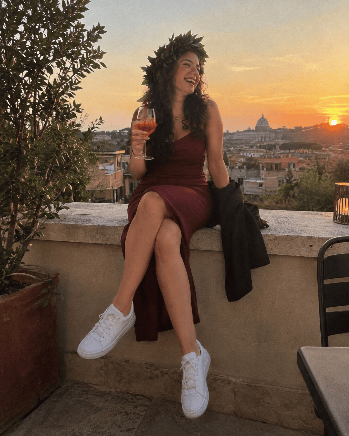 Laureanda con abito midi bordeaux, corona d'alloro e sneaker bianche minimal alla festa di laurea su una terrazza al tramonto, look per il dopo proclamazione