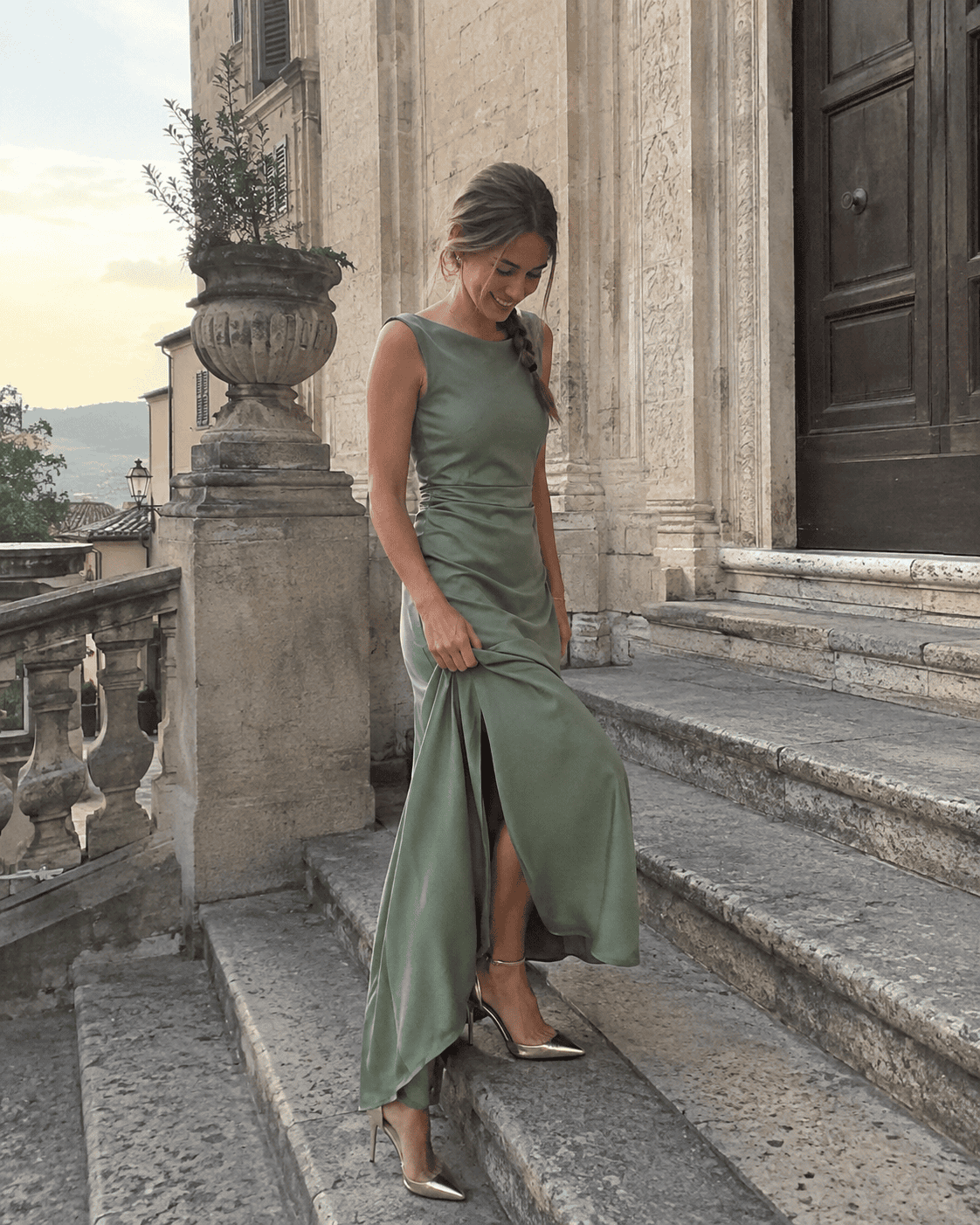 Laureanda con abito lungo verde salvia in tessuto opaco con spacco discreto e décolleté dorate sulla scalinata di un palazzo storico, outfit laurea elegante
