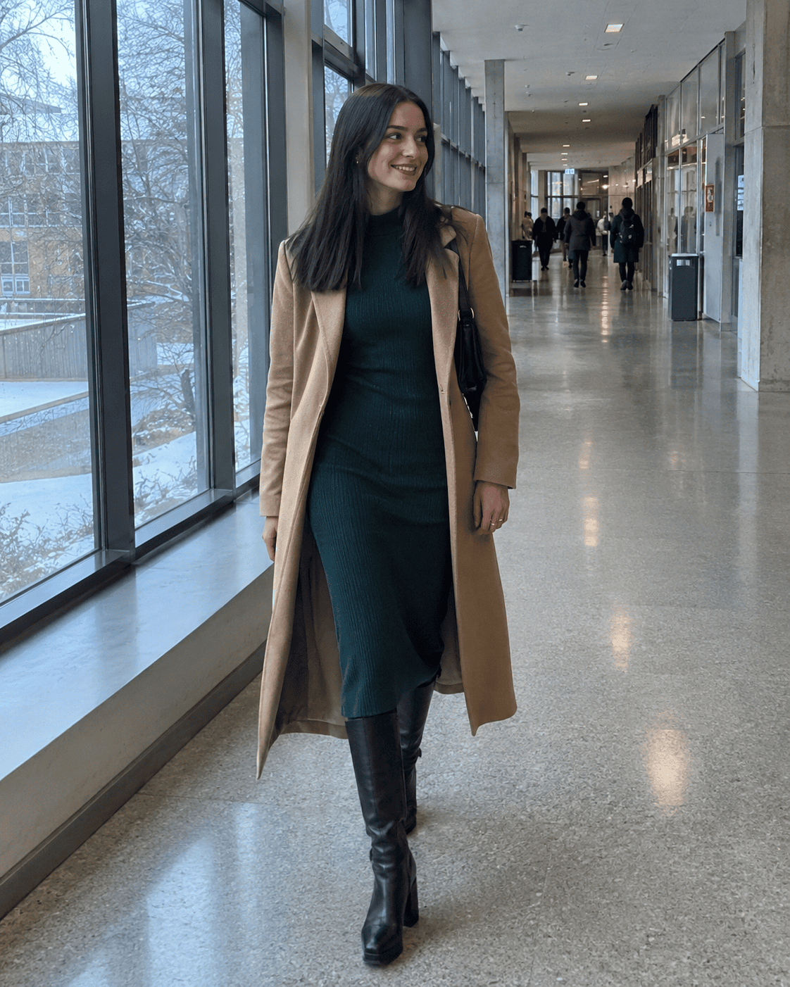 Laureanda con abito midi in maglia verde bottiglia, stivali alti neri e cappotto lungo cammello nel corridoio dell'università, outfit laurea inverno