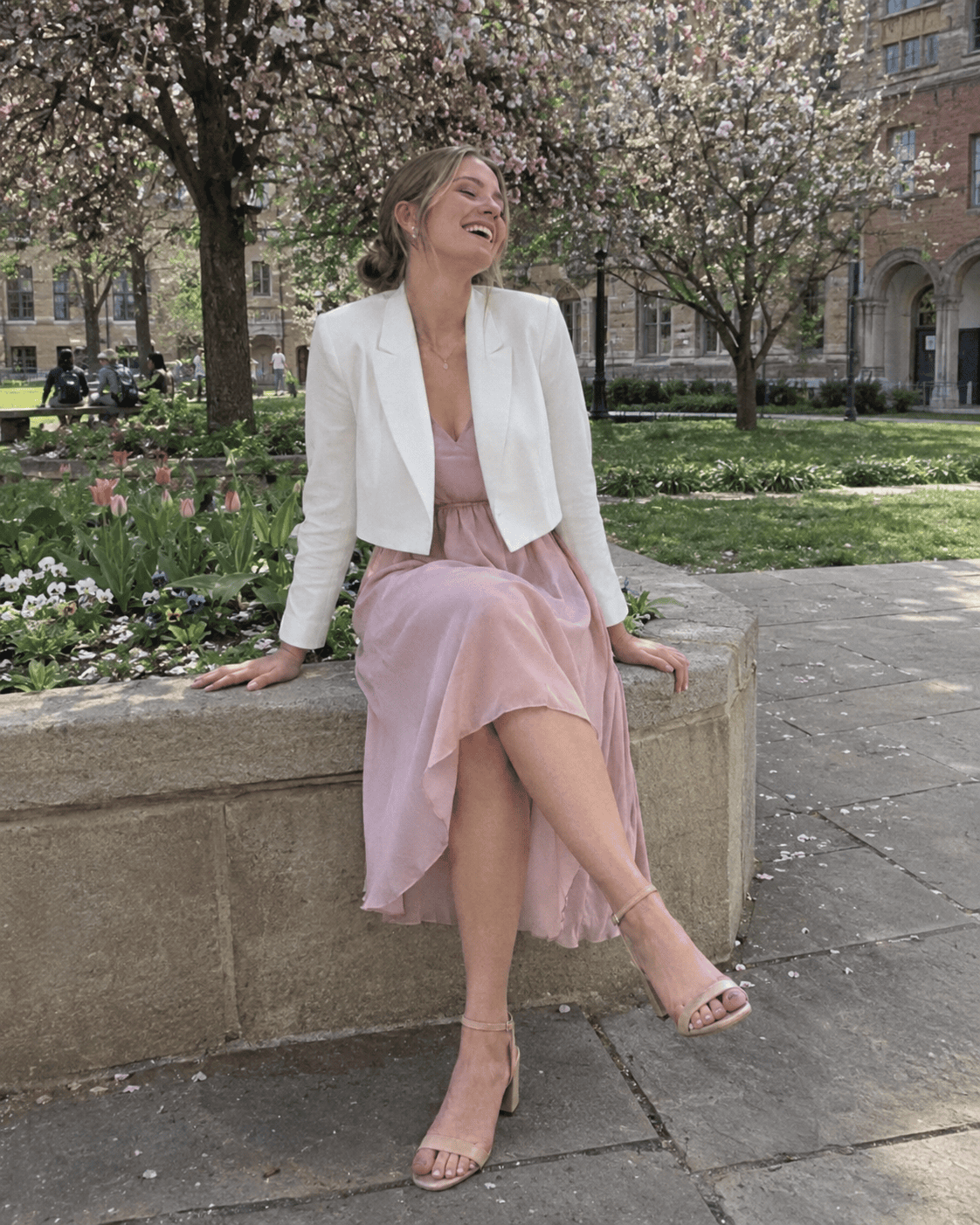 Laureanda con abito fluido rosa cipria, blazer corto bianco e sandali nude con tacco medio nel cortile fiorito dell'università, outfit laurea primavera