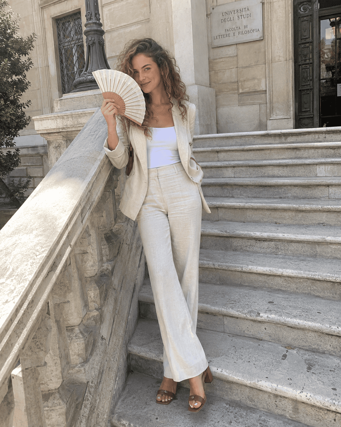 Laureanda con completo in lino color avorio, top bianco, sandali cuoio con tacco medio e ventaglio sulla scalinata dell'università, outfit laurea estate