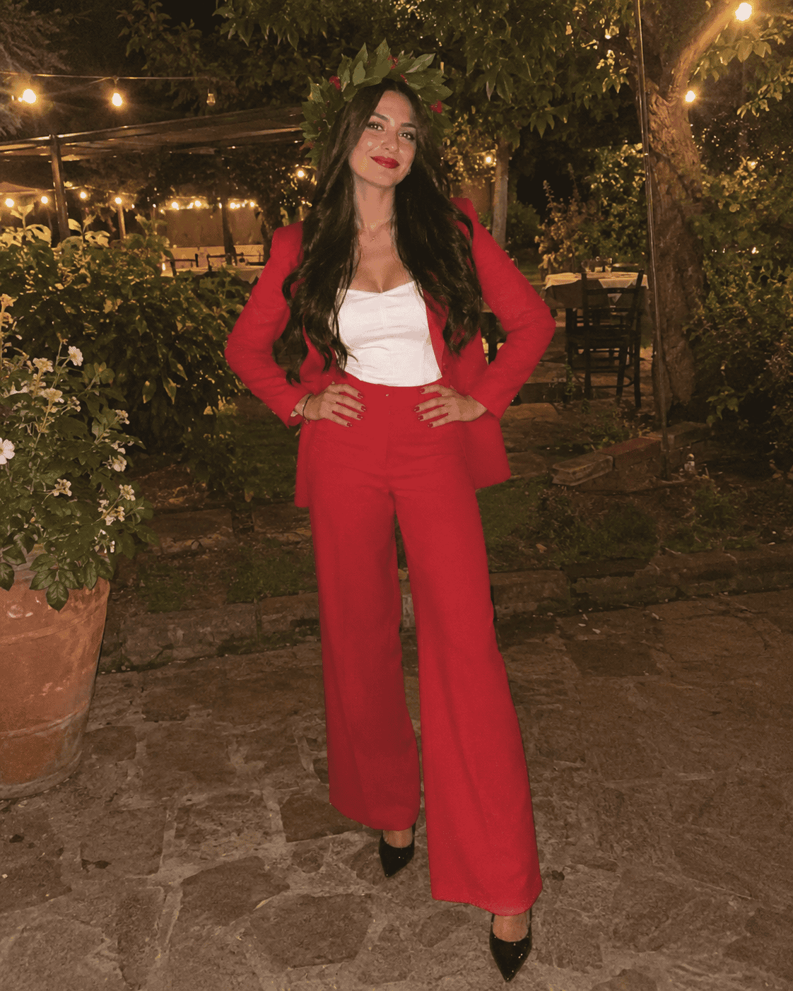 Laureanda con completo rosso giacca e pantalone palazzo, top bianco, décolleté nere e corona d'alloro alla festa di laurea in giardino, outfit laurea