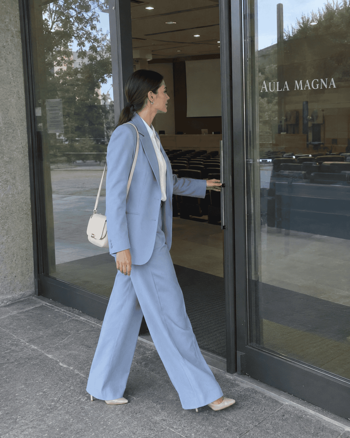 Laureanda con tailleur azzurro polvere, camicia di seta bianca, décolleté nude e borsa crema all'ingresso dell'aula magna, outfit laurea elegante