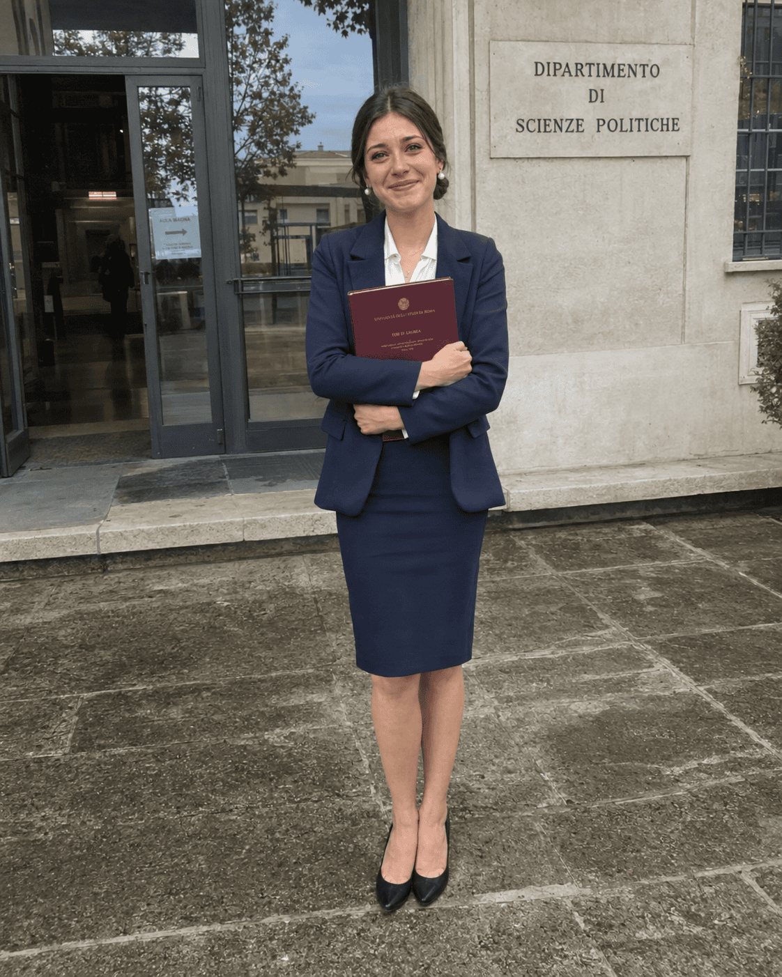 Laureanda con tailleur blu navy, gonna a tubino, camicia bianca, orecchini a perla e tesi bordeaux tra le braccia davanti al dipartimento, outfit laurea magistrale