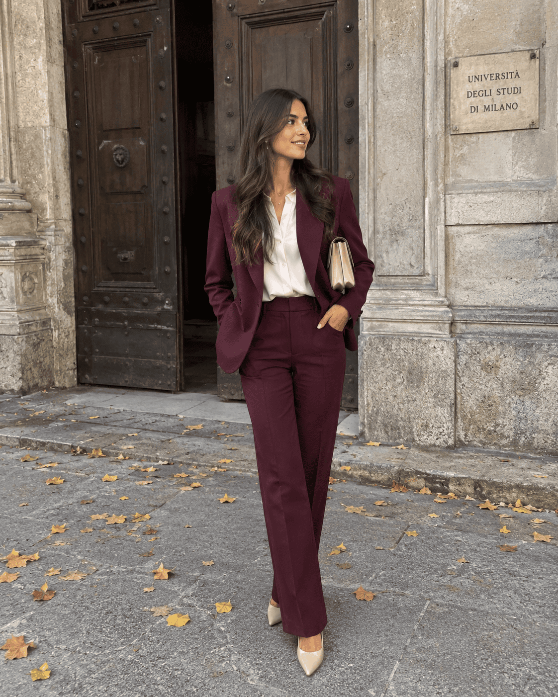 Laureanda in tailleur bordeaux con camicia panna, décolleté nude e pochette beige davanti al portone dell'università, outfit laurea autunno