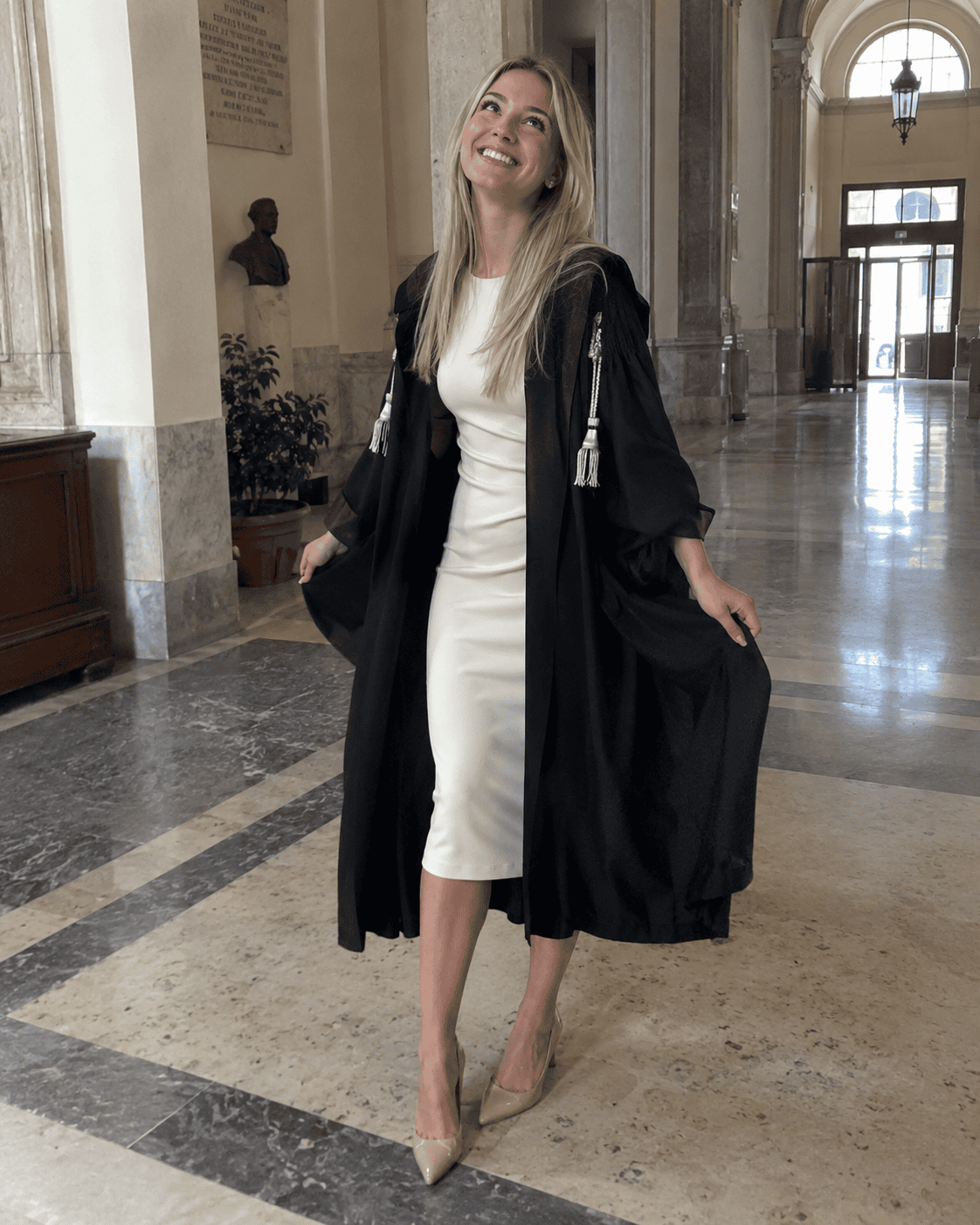 Laureanda con toga nera aperta sopra un abito avorio al polpaccio e décolleté nude nell'atrio dell'università, cosa mettere sotto la toga